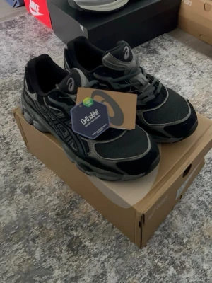 Svarta Asics sneakers  - Snygga svarta Asics sneakers med grå detaljer. Ovandelen är i mesh och syntetiskt skinn för bra andningsförmåga och stöd. Klassisk Asics-logga på sidan och Ortholite-innersula för extra komfort. Perfekta för dig som gillar sportig och stilren look.