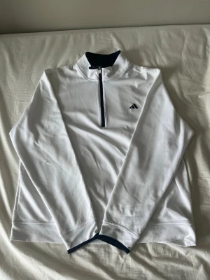 Vit långärmad träningströja Adidas - Snygg vit långärmad golf tröja från Adidas med halv dragkedja framtill och diskret logga på bröstet. Tröjan har mörkblå detaljer vid krage och ärmslut. Tillverkad i ett lätt och funktionellt material som passar perfekt till träning eller uppvärmning.