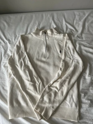 Beige stickad halvzip från Ralph Lauren - Klassisk vit stickad tröja från Ralph Lauren med halvzip och hög krage. Tröjan har långa ärmar, ribbade muddar och den ikoniska lilla loggan broderad på bröstet. Perfekt till jeans eller chinos för en clean och stilren look.