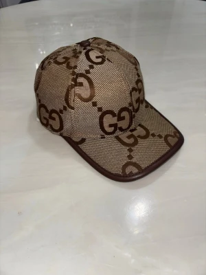 Beige keps med GG-mönster från Gucci - Snygg beige keps från Gucci med klassiskt GG-monogram i brunt över hela kepsen. Justerbar rem baktill och böjd skärm. Materialet är textil med läderdetaljer längs kanten. Perfekt för dig som vill sticka ut med en lyxig accessoar.
