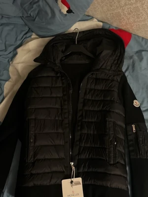 Svart pufferjacka från Moncler med huva - Snygg svart pufferjacka från Moncler med huva och dragkedja framtill. Jackan har quiltad front och rygg, samt stickade ärmar med Moncler-logga på ena ärmen och en praktisk ficka med dragkedja. Perfekt för kyliga dagar.