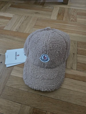 Beige teddykeps från Moncler - Säljer en beige keps från Moncler i mjukt teddy-material. Kepsen har böjd skärm och ett broderat Moncler-märke framtill. Perfekt för dig som vill ha en trendig och mysig accessoar till hösten eller vintern.