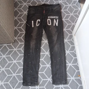 Svarta Dsquared2 ICON jeans med kedja - Snygga svarta jeans från Dsquared2 med stor vit 'ICON'-print på baksidan och coola slitningar samt färgstänk. Kedjedetalj med Dsquared2-mynt vid fickan ger extra edge. Smal passform och klassisk femficksdesign. Perfekta för dig som vill sticka ut.