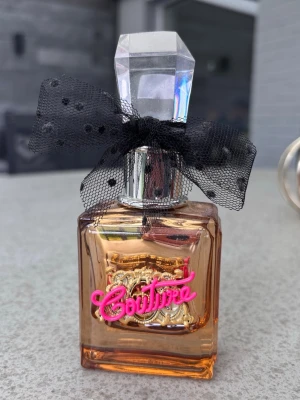 Juicy Couture parfym  - Juicy Couture parfym i en fyrkantig glasflaska med guld- och rosa detaljer samt texten 'Couture' i rosa framtill. Flaskan har en stor, kristalliknande kork och en svart prickig rosett runt halsen. Perfekt för dig som gillar lyxig och lekfull design.