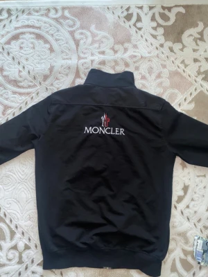 Svart Moncler  - Snygg svart jacka från Moncler med hög krage och dragkedja framtill. Jackan har broderad Moncler-logga på bröstet och stor logotyp på ryggen. Tillverkad i ett lätt material som passar perfekt till streetwear och vardagsstil.