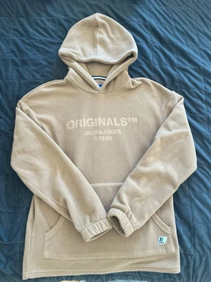 Beige fleecehoodie Jack & Jones - Mysig beige hoodie i fleece från Jack & Jones med broderad text 'ORIGINALS™ JACK & JONES ©1990' på bröstet. Hoodien har huva, känguruficka och elastiska muddar vid ärmsluten. Perfekt för chill dagar eller när du vill vara extra bekväm.