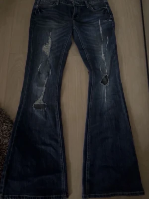 Mörkblå utsvängda jeans med slitningar - Säljer ett par mörkblå jeans med utsvängda ben och snygga slitningar framtill. Jeansen har klassisk femficksdesign och låg midja. Perfekta för dig som gillar en avslappnad och trendig look.