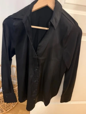 Svart satinliknande skjorta - Stilren svart skjorta i ett glansigt, satinliknande material. Skjortan har klassisk krage, långa ärmar och knäppning framtill. Perfekt för dig som gillar en clean och modern look.