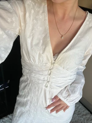 Vit långklänning med broderade detaljer - Romantisk vit långklänning med broderade blommor, v-ringning och knappar framtill. Klänningen har långa ärmar och markerad midja för en smickrande siluett. Perfekt för sommarens speciella tillfällen.