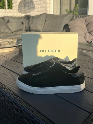 Svarta sneakers från Axel Arigato - Snygga svarta sneakers från Axel Arigato med dragkedja framtill och vit sula. Tillverkade i mocka och läder för en clean och modern look. Perfekta för dig som gillar minimalistisk stil och vill ha något som sticker ut lite extra.
