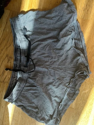 Randiga svarta shorts med snörning - Säljer ett par svarta shorts med tunna vita ränder och snörning i midjan. De har en avslappnad passform och är perfekta för varma dagar. Shortsen är tillverkade i ett mjukt och lätt material som känns skönt mot huden.