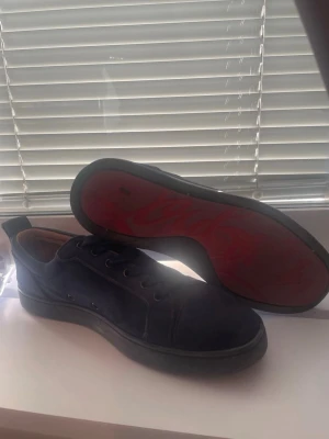 Loubs - Snygga mörkblå sneakers i mocka med klassisk låg profil och svarta skosnören. Skorna har en unik röd sula och diskreta metalldetaljer på sidan. Insidan är fodrad i beige läder för extra komfort. Perfekta för dig som gillar stilrena och exklusiva sneakers.