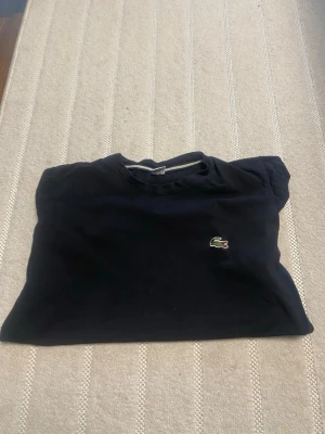 Svart t-shirt från Lacoste - Klassisk svart t-shirt från Lacoste med den ikoniska krokodilloggan broderad på bröstet. T-shirten har rund halsringning och korta ärmar. Perfekt basic-plagg med stilren design och skön passform.
