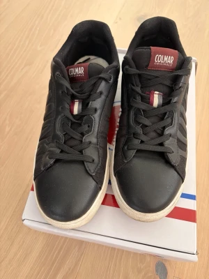 Svarta Colmar Originals sneakers - Snygga svarta sneakers från Colmar Originals med vita sulor och detaljer i rött och vitt på plösen. Skorna har klassisk låg modell, svarta snören och ovandel i en mix av textil och syntet. Perfekta för en stilren och sportig look.