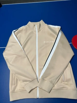 Beige Palm Angels hoodie med dragkedja - Snygg beige hoodie från Palm Angels med vit dragkedja och vita ränder längs ärmarna. Märkets logga i vitt på bröstet. Tillverkad i ett mjukt och stretchigt material, perfekt för en avslappnad streetwear-look.