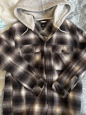 Rutig overshirt med huva från H&M - Säljer en snygg overshirt från H&M i brun- och svartvitrutigt mönster. Skjortjackan har två bröstfickor med knappar och en grå huva i sweatshirtmaterial med dragsko. Perfekt lager på lager-plagg för en avslappnad stil.