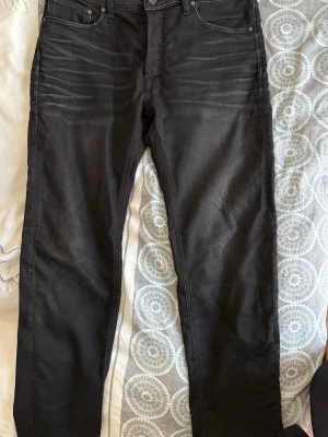 Svarta jeans från Jack & Jones - Säljer ett par svarta jeans från Jack & Jones med klassisk femficksmodell och smal passform. Jeansen har snygga slitningar vid låren och är tillverkade i stretchigt material för extra komfort. Perfekta för en avslappnad och trendig look.
