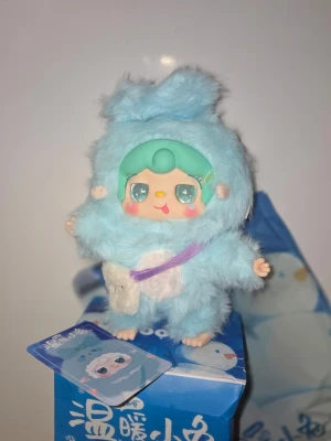 Söt blå YOOKI  samlarfigur med fluffig dräkt ur Warm bunny serien - NY Samlarfigur YOOKI ur warm bunny serien höjd ca 23 - 24 cm med mintgrönt hår och stora glittriga ögon, klädd i en fluffig ljusblå dräkt med öron och vit mage. Figurer har en liten lila axelväska och står på en blå kartong med matchande illustration. Perfekt för dig som gillar gulliga prydnader. 
