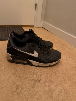 Svarta Nike Air Max sneakers - Snygga svarta Nike Air Max sneakers med vit sula och vit swoosh på sidan. Skorna har en klassisk låg siluett, mesh- och syntetmaterial samt synlig Air-enhet i hälen för extra komfort. Perfekta för dig som gillar streetwear och sportig stil.