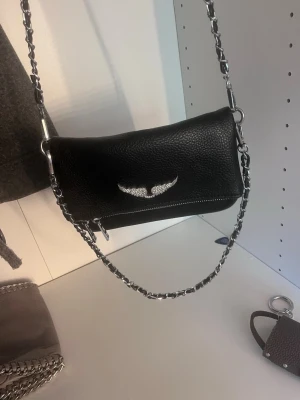 Svart axelväska med kedjerem - Snygg svart axelväska i präg­lat läder med silverfärgad kedjerem och dragkedja baktill. Framsidan har en cool silverdetalj i form av vingar. Perfekt för dig som vill ha en edgy och trendig look.