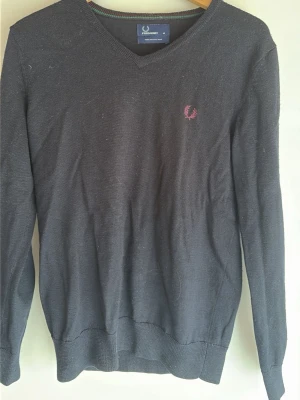 Svart V-ringad tröja Fred Perry stickat  - Klassisk svart V-ringad tröja från Fred Perry med den ikoniska vinröda lagerkransen broderad på bröstet. Tröjan har långa ärmar och ribbade muddar vid ärmslut och nederkant. Perfekt för en stilren och enkel look.