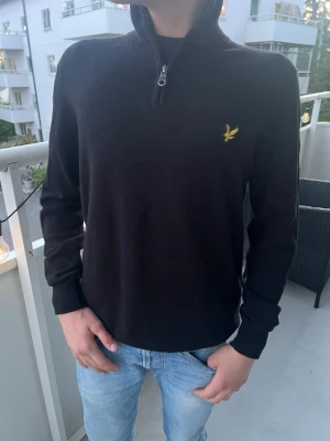 Svart stickad tröja från Lyle & Scott - Svart stickad tröja från Lyle & Scott med halv dragkedja och gul logga på bröstet. Tröjan har lång ärm, ribbade muddar och klassisk passform. Perfekt till jeans för en stilren look