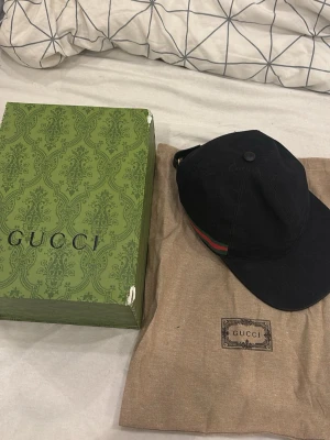 Svart keps från Gucci med röd/grön detalj - Säljer en svart keps från Gucci med klassisk röd och grön rand på sidan. Kepsen är tillverkad i Italien och har justerbar rem bak. Materialet är bomull och kepsen kommer med originalkartong och dustbag.