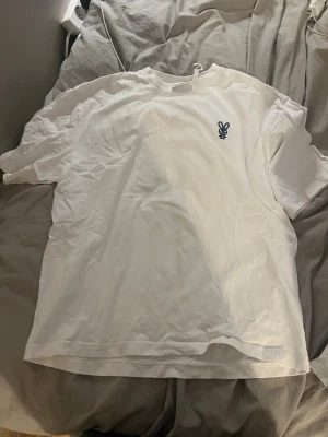 Vit t-shirt från Weekday med blå logga - Vit t-shirt från Weekday i bomull med en liten blå broderad logga på bröstet. Klassisk rund hals och korta ärmar, perfekt för en clean och enkel stil.