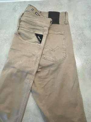Beige jeans från Replay - Snygga beige jeans från Replay med klassisk femficksdesign och diskret broderad detalj på bakfickan. Jeansen har en rak passform och är tillverkade i ett mjukt bomullsmaterial med lite stretch för extra komfort. Ny tvättade 