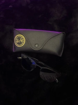 Svarta Ray-Ban solglasögon - Snygga svarta solglasögon från Ray-Ban med klassisk bågform och diskret logga på skalmen. Kommer med original svart fodral i läderlook. Perfekta för att lyfta din stil under soliga dagar.