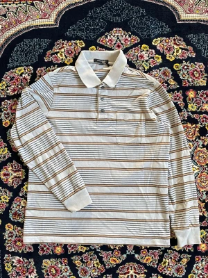 Randig långärmad polotopp - Säljer en långärmad polotopp från Weekday med tunna horisontella ränder i beige, brunt och svart. Toppen har en klassisk krage, knäppning framtill och en bröstficka. Perfekt för dig som gillar en avslappnad men stilren look.