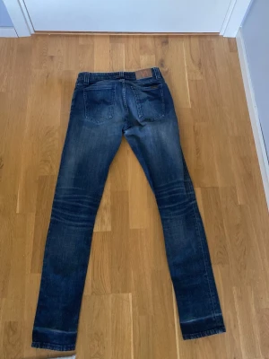 Mörkblå skinny jeans från Nudie - Snygga mörkblå slim jim  jeans från Nudie med klassiska bakfickor och diskreta slitningar. Jeansen har en smal passform och är tillverkade i stretchig denim för extra komfort. Perfekta för en trendig och avslappnad stil.