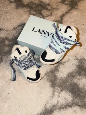 Lanvin vita och svarta high top sneakers - Lanvin high top sneakers i vitt och svart med detaljer i ljusblått och mintgrönt. Skorna har breda, mönstrade skosnören i blått och vitt samt en chunky sula. Tillverkade i läder och mocka med tydliga kontrastpaneler och logga på plösen. ALDRIG ANVÄNDA.