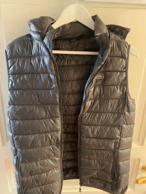 Grå dunväst från Lacoste med huva - Snygg grå dunväst från Lacoste med quiltad design och avtagbar huva. Västen har dragkedja framtill, två fickor med dragkedja och den klassiska Lacoste-loggan på bröstet. Perfekt för lager-på-lager och chill stil. Kan ta av luvan.