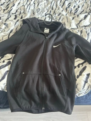 Svart Nike zip hoodie med huva - Säljer en svart hoodie från Nike med dragkedja framtill och klassisk vit Swoosh-logga på bröstet. Hoodien har huva, två fickor fram och är tillverkad i mjukt bomullsmaterial. Perfekt för en avslappnad och sportig stil. Storlek M men passar S. Den har använts men har inga tecken på skador