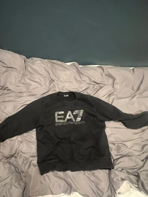 Svart EA7 Emporio Armani sweatshirt - Snygg svart sweatshirt från EA7 Emporio Armani med stor logga i silver på bröstet. Tröjan har rund halsringning och långa ärmar. Perfekt för dig som gillar stilrena och exklusiva plagg.