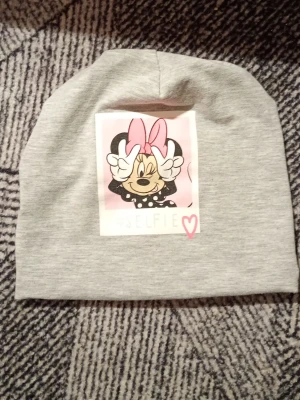 Grå mössa med Minnie Mouse-tryck - Snygg grå mössa med ett stort tryck av Minnie Mouse som gör en selfie-pose. Trycket har rosa och svarta detaljer samt texten #SELFIE med ett litet hjärta. Perfekt accessoar för dig som gillar Disney och vill ha en lekfull stil.