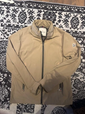 Beige vindjacka från Moncler - Stilren beige vindjacka från Moncler med hög krage och dragkedja framtill. Jackan har två fickor med dragkedja och Moncler-logga på ena ärmen. Perfekt för dig som gillar en clean och sportig look.
