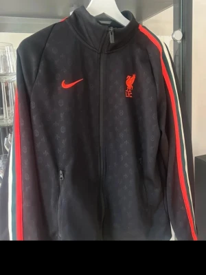 Liverpool Nike svart träningsjacka - Svart träningsjacka från Nike med Liverpool FC-logga i rött på bröstet. Jackan har hel dragkedja, röda och vita ränder längs ärmarna och diskret mönster över hela tyget. Perfekt för fotbollsfans som vill ha en sportig look. Nypris 899