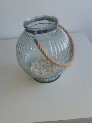 Rund glaslykta med rep - Säljer en rund glaslykta med räfflad yta och ett grovt rep som handtag. Perfekt som dekoration eller för att ha värmeljus i. Glaset är genomskinligt och handtaget är i naturfärgat rep. Metallkant upptill för extra stabilitet.
