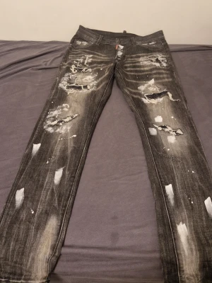 Svarta slitna jeans från DSQ2 Denim - Riktigt snygga svarta jeans från DSQ2 Denim med slitningar, hål och vitmålade detaljer. Jeansen har raka ben och klassisk femficksdesign. Perfekta för dig som gillar en edgy och trendig stil.