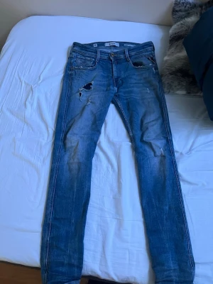 Blå slitna jeans från Replay - Säljer ett par blå jeans från Replay med slitningar och hål på låret. Klassisk femficksmodell med raka ben och låg midja. Jeansen har en snygg tvättad look och detaljerade sömmar. Perfekta för en avslappnad och trendig stil.