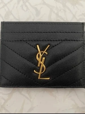 Svart ysl plånbok  - Snyggt svart kortfodral från Yves Saint Laurent i präglat läder med guldfärgad YSL-logga framtill. Fodralet har flera kortfack och elegant chevronmönster. Perfekt för att hålla koll på dina kort med stil.