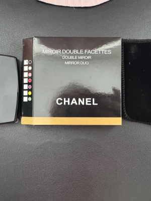 Chanel Miroir Double Facettes sminkspegel - Snygg och stilren sminkspegel från Chanel, modell Miroir Double Facettes. Spegeln är svart med klassisk Chanel-logga och kommer i en elegant förpackning samt ett svart sammetsfodral. Perfekt storlek för att ha i väskan och enkel att ta med överallt.