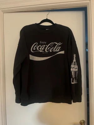 Gråsvart Coca-Cola sweatshirt  - Cool gråsvart sweatshirt med stort Coca-Cola-tryck på bröstet och en klassisk Coca-Cola-flaska tryckt på ena ärmen. Tröjan har rund hals och ribbade muddar vid ärmslut och nederkant. 