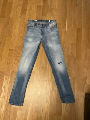 Jack & Jones jeans slim fit glenn - Säljer ett par ljusblå jeans med slitning på ena benet och snygga detaljer. Jeansen har fem fickor, knapp och dragkedja framtill samt stretchigt material för skön passform. Perfekt för dig som gillar en avslappnad och trendig look. Storlek: 164 