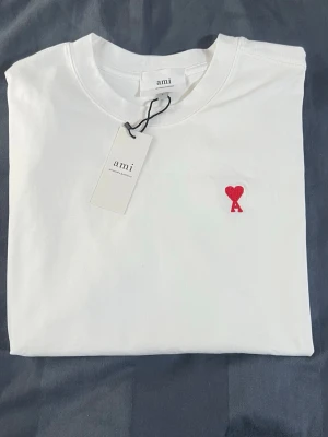Vit t-shirt från Ami med hjärtbroderi - Vit t-shirt från Ami med ett snyggt rött broderat hjärta och bokstaven A på bröstet. Klassisk rund halsringning och korta ärmar. Gjord i mjuk bomull som känns skön mot huden. Perfekt för dig som gillar stilrena plagg med en twist.