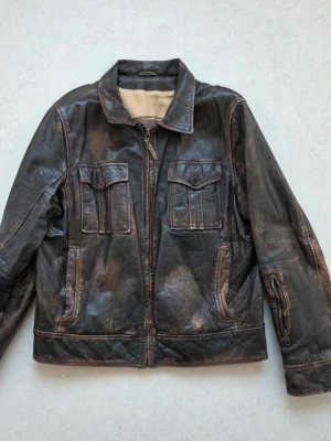 Brun vintage skinnjacka med fickor - Cool brun vintage skinnjacka med dragkedja framtill och två bröstfickor med lock och knapp. Klassisk krage och detaljerade sömmar ger jackan en retro känsla. Perfekt för dig som gillar en tidlös och avslappnad stil.