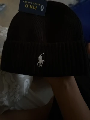 Svart ribbstickad mössa Polo Ralph Lauren - Snygg svart ribbstickad mössa från Polo Ralph Lauren med klassiska loggan broderad framtill. Mössan har uppvikt kant och är tillverkad i mjukt material som håller dig varm. Perfekt accessoar för kalla dagar och enkel att matcha med olika outfits.