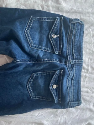 Mörkblå bootcut jeans  - Säljer ett par mörkblå bootcut jeans med vita kontrastsömmar och snygga ficklock bak med silverknappar. Jeansen har klassisk femficksmodell och är tillverkade i stretchigt denimtyg för skön passform. Perfekta för en trendig och avslappnad look.